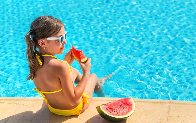 Menina de óculos e roupa de banho comendo melancia na beirada da piscina, em dúvida se deve nadar em seguida.