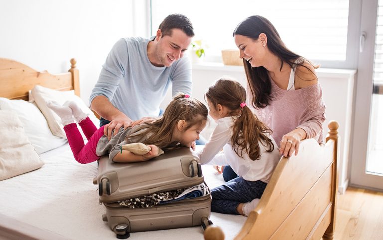Família composta por pai, mãe e duas filhas pequenas preparando a mala para a primeira viagem das crianças.