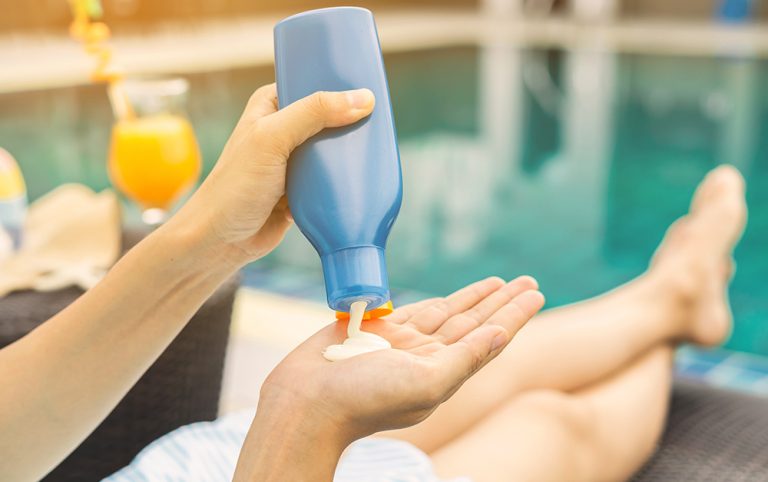 Protetor solar sendo aplicado em uma mão, com piscina em fundo embaçado.