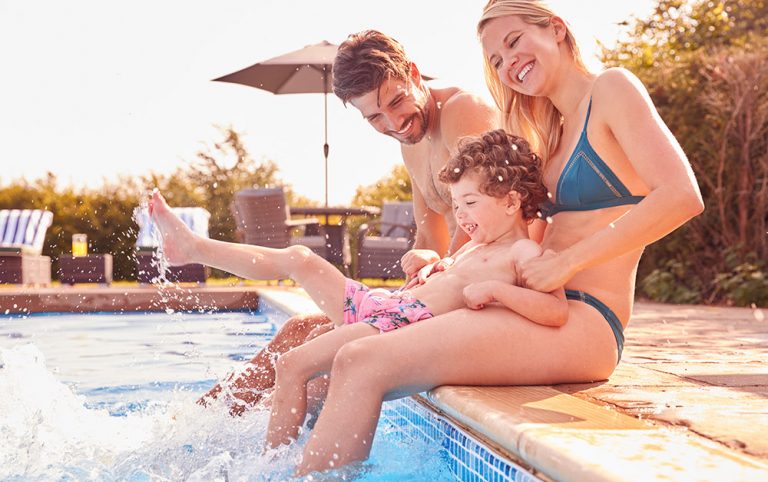 Família composta por pai, mãe e filho pequeno sentados fazendo brincadeiras à beira de uma piscina.