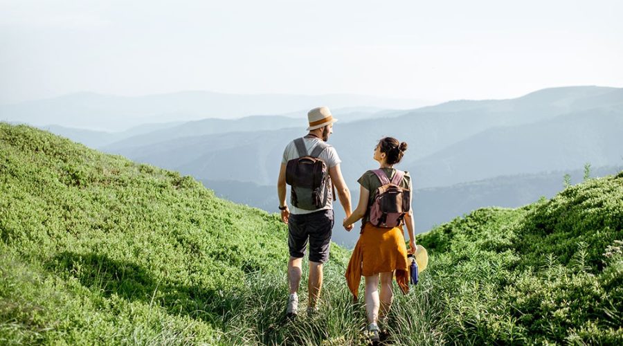 Viagem de casal, onde ambos exploram uma região serrana enquanto caminham de mãos dadas e mochila nas costas