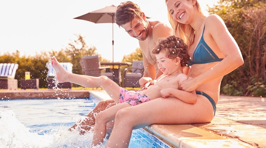 Família composta por pai, mãe e filho pequeno sentados fazendo brincadeiras à beira de uma piscina.