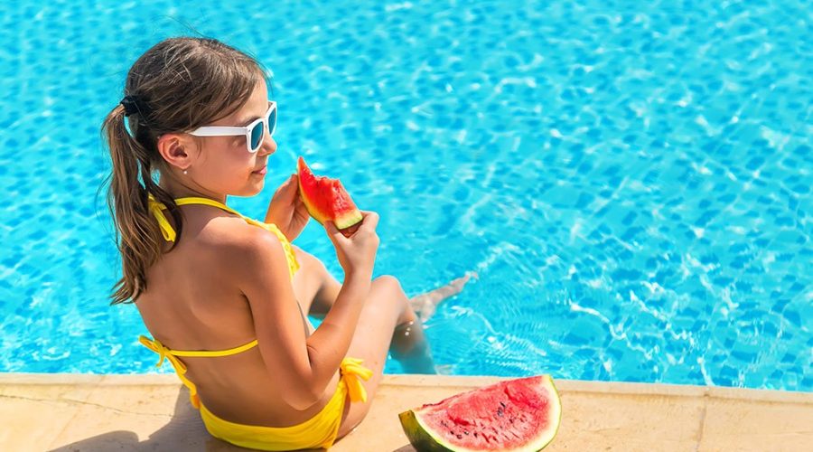 Menina de óculos e roupa de banho comendo melancia na beirada da piscina, em dúvida se deve nadar em seguida.