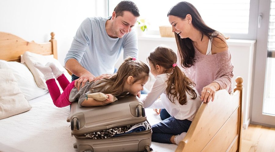 Família composta por pai, mãe e duas filhas pequenas preparando a mala para a primeira viagem das crianças.